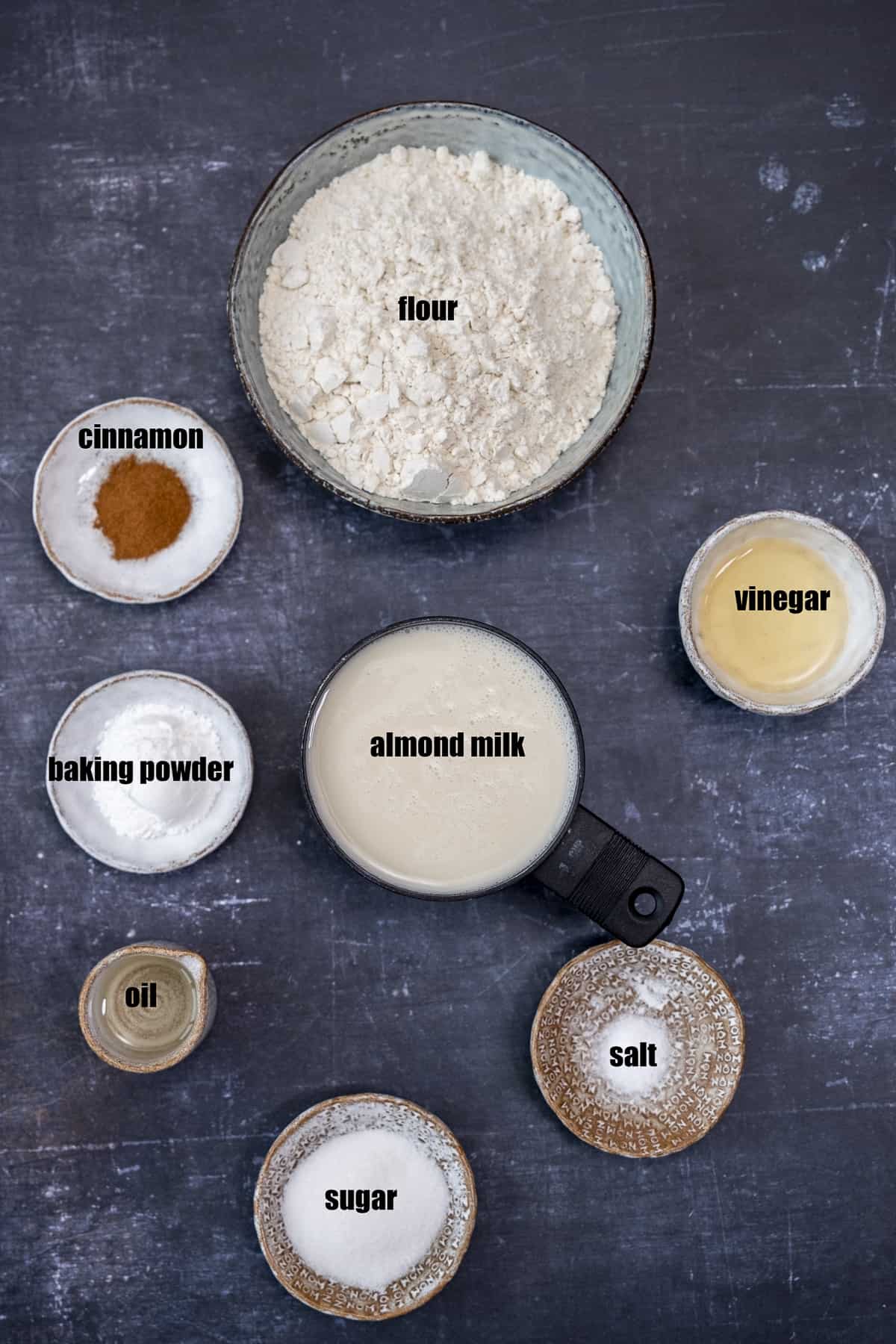 Almond milk, vinegar, oil, flour, cinnamon, sugar, salt and baking powder in separate bowls on a dark background.
