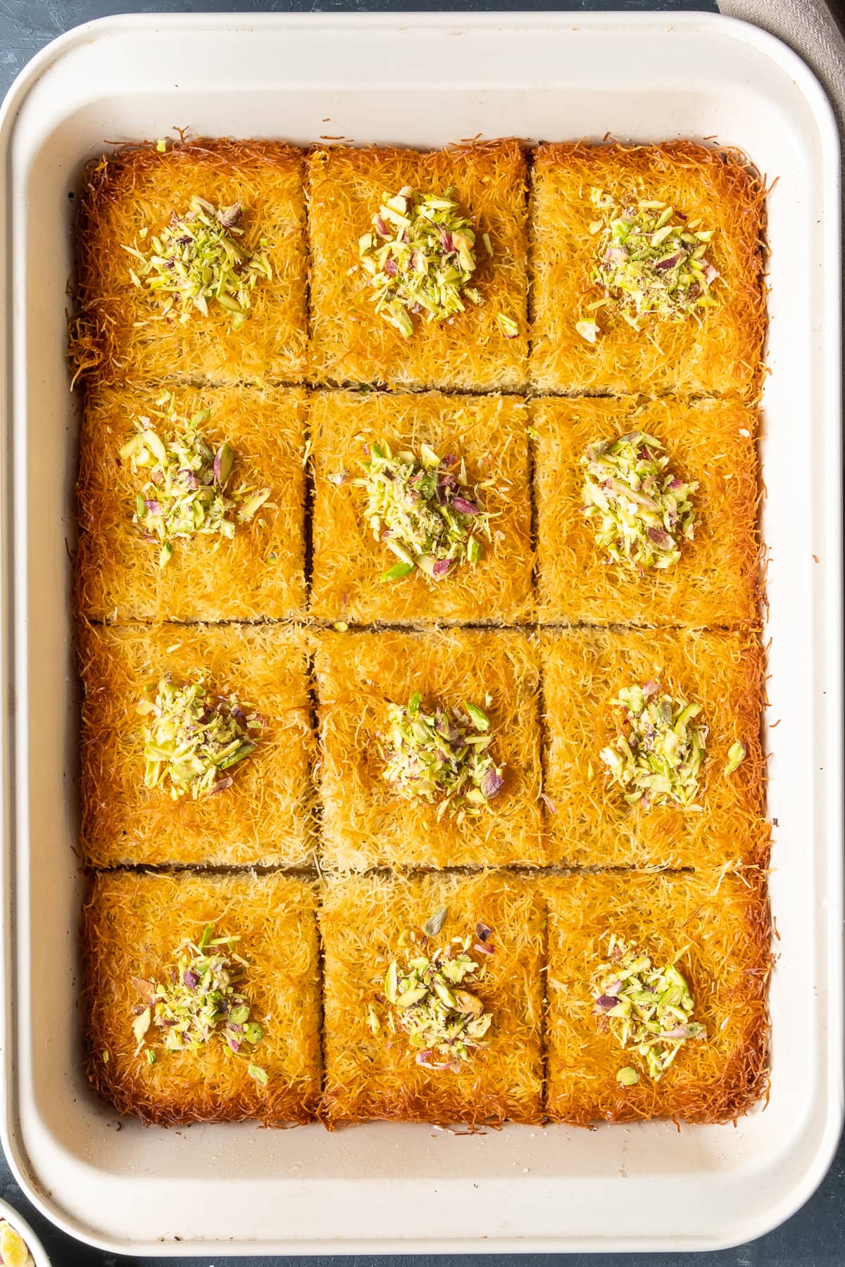 Golden and crispy kadaif dessert sliced in squares and topped with crumbled pistachios in a rectangular pan.