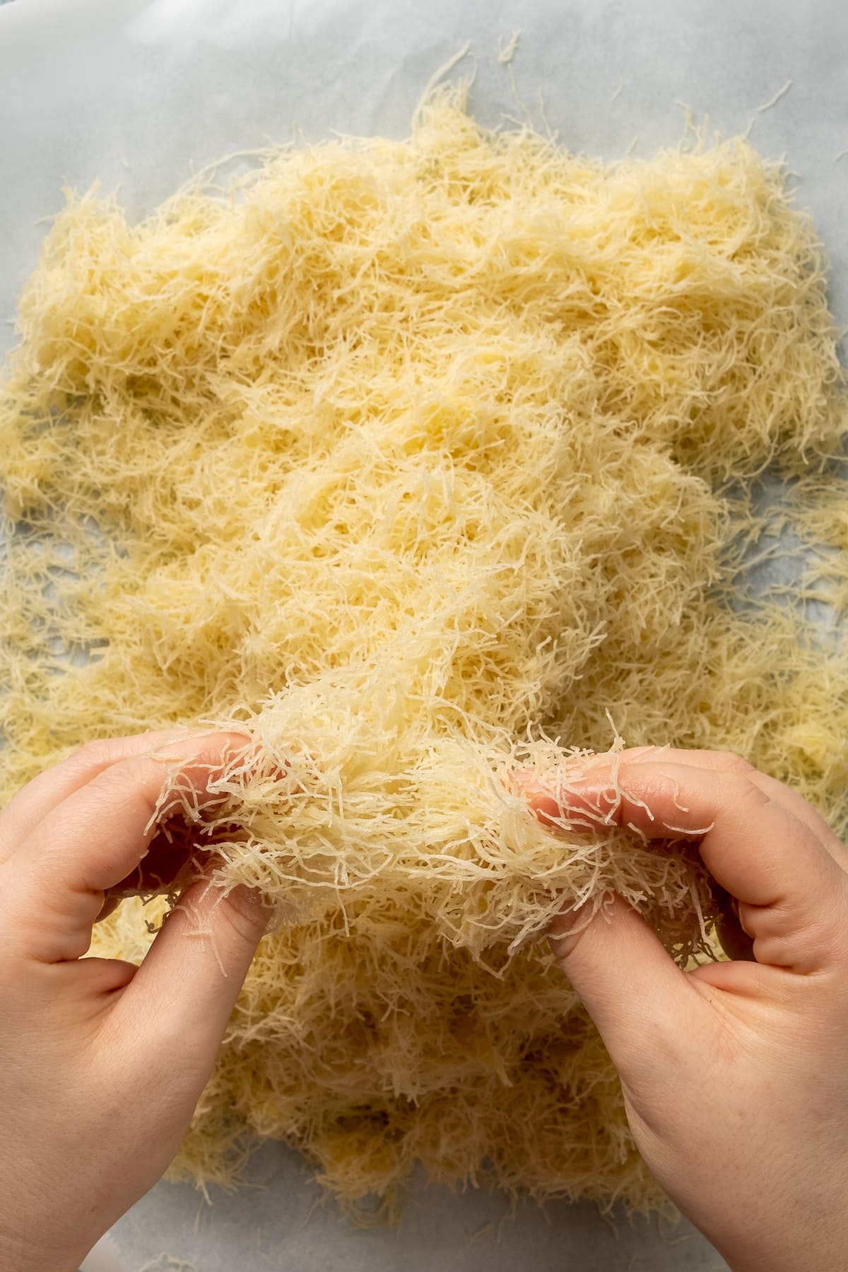 Hands coating the kadaif noodles with melted butter on a piece of baking paper.