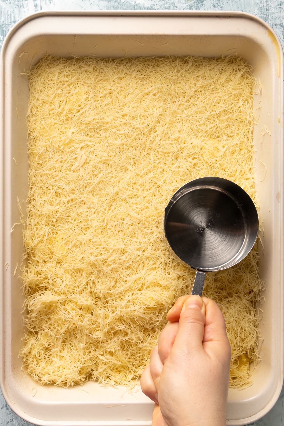 A hand flattening shredded and buttered kadayif noodles in a rectangular baking pan.