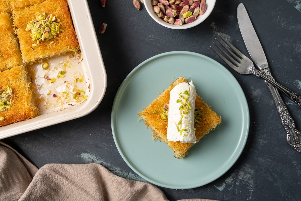 A slice of kadaif topped with a generous roll of clotted cream aka kaymak on a blueish plate, whole pistachios in a bowl and the rest of kadaif dessert in the pan on the side.