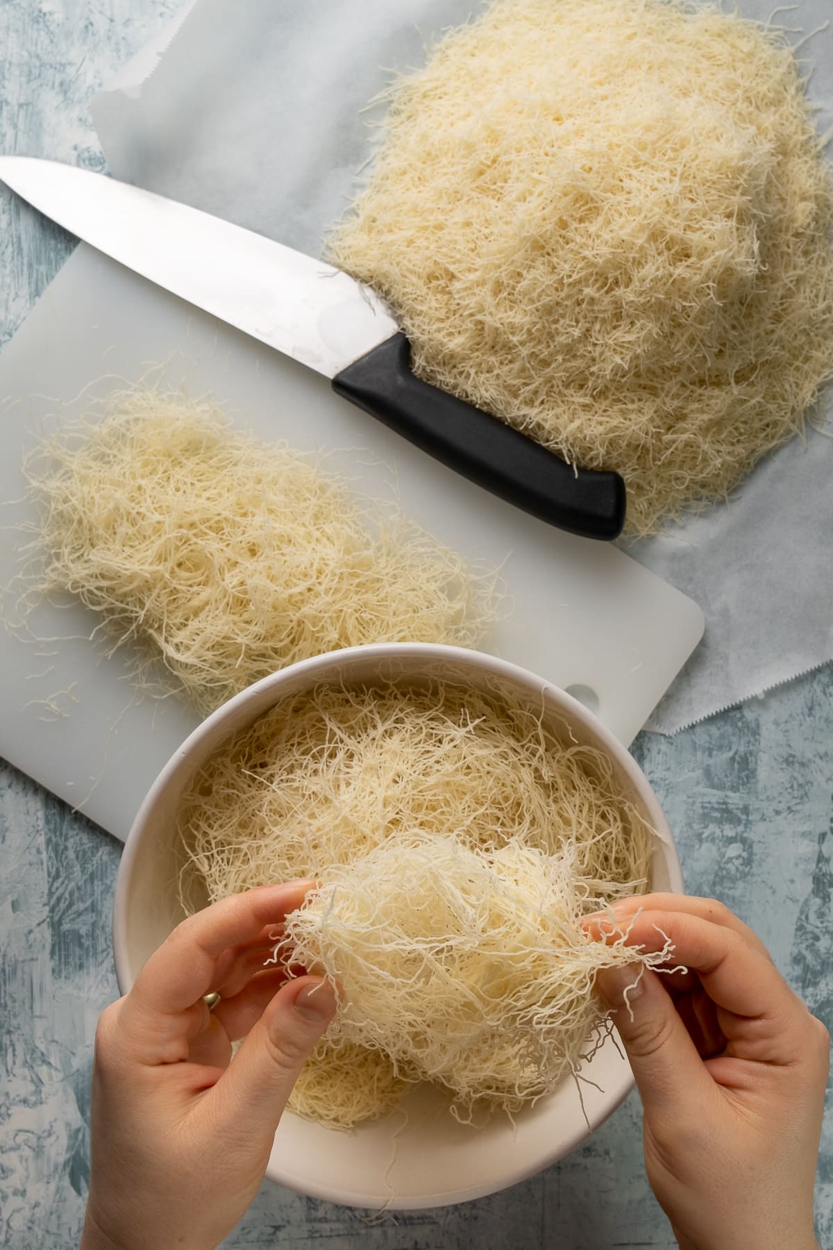 Hands shredding kadaif noodles in a bowl, more noodles on a cutting board and on a piece of baking paper and a knife on the ground.