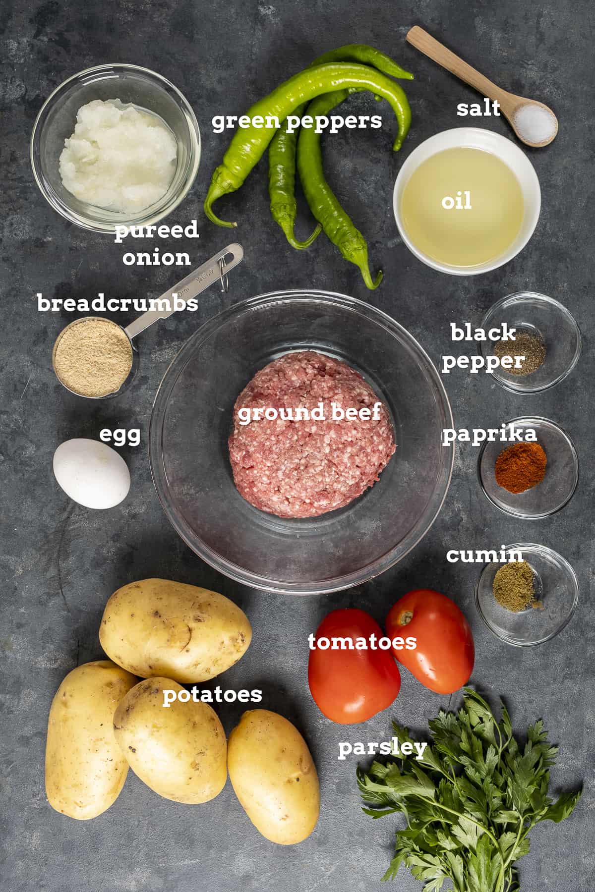 Ground beef, potatoes, tomatoes, parsley, pureed onion, oil, breadcrumbs and spices on a dark background.