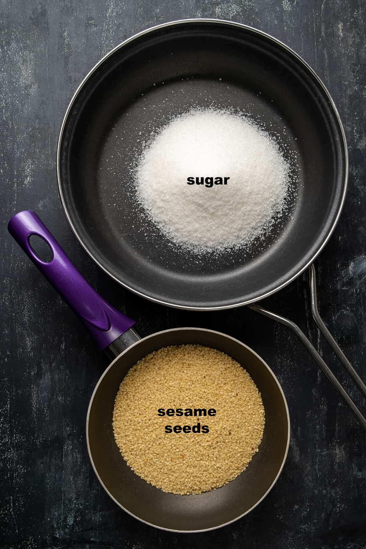 Granulated sugar in a non stick pan and raw sesame seeds in a smaller pan next to it.