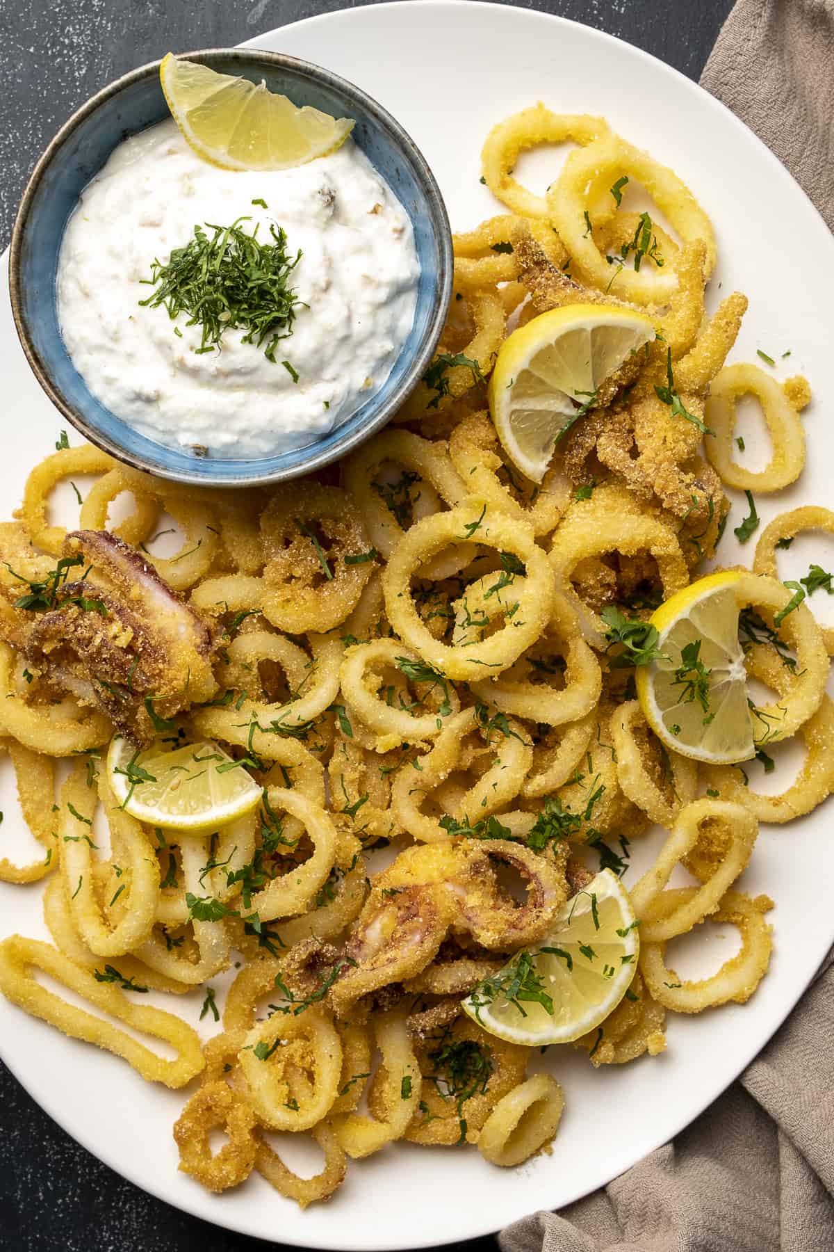 Fried calamari rings with lemon slices on a white plate. A mayo sauce in a small bowl on the side.
