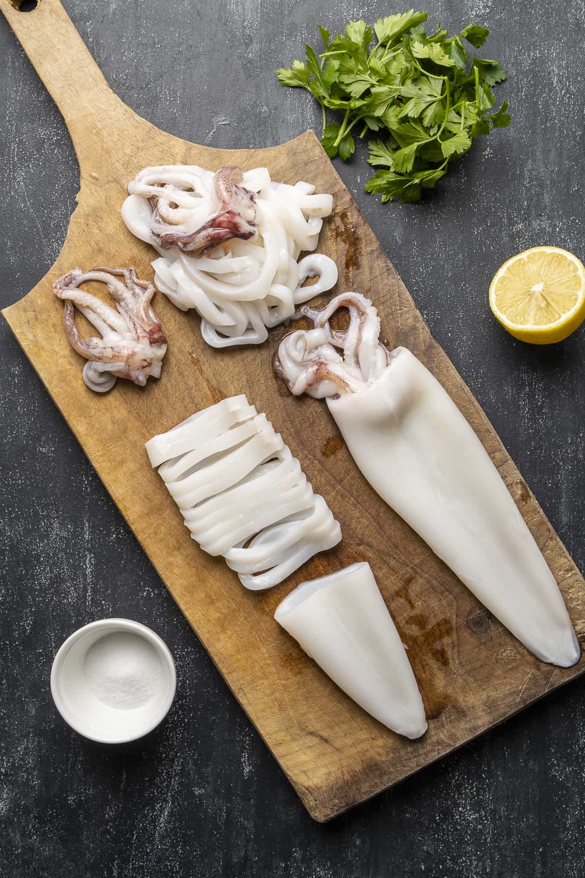 Whole squids on a cutting board, partly sliced, parsley and lemon on the ground.