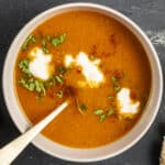 Carrot and tomato soup topped with yogurt, parsley and red pepper flakes in a white bowl and a spoon in it.