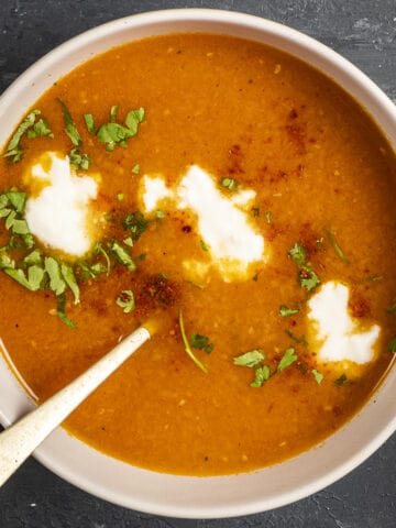 Carrot and tomato soup topped with yogurt, parsley and red pepper flakes in a white bowl and a spoon in it.