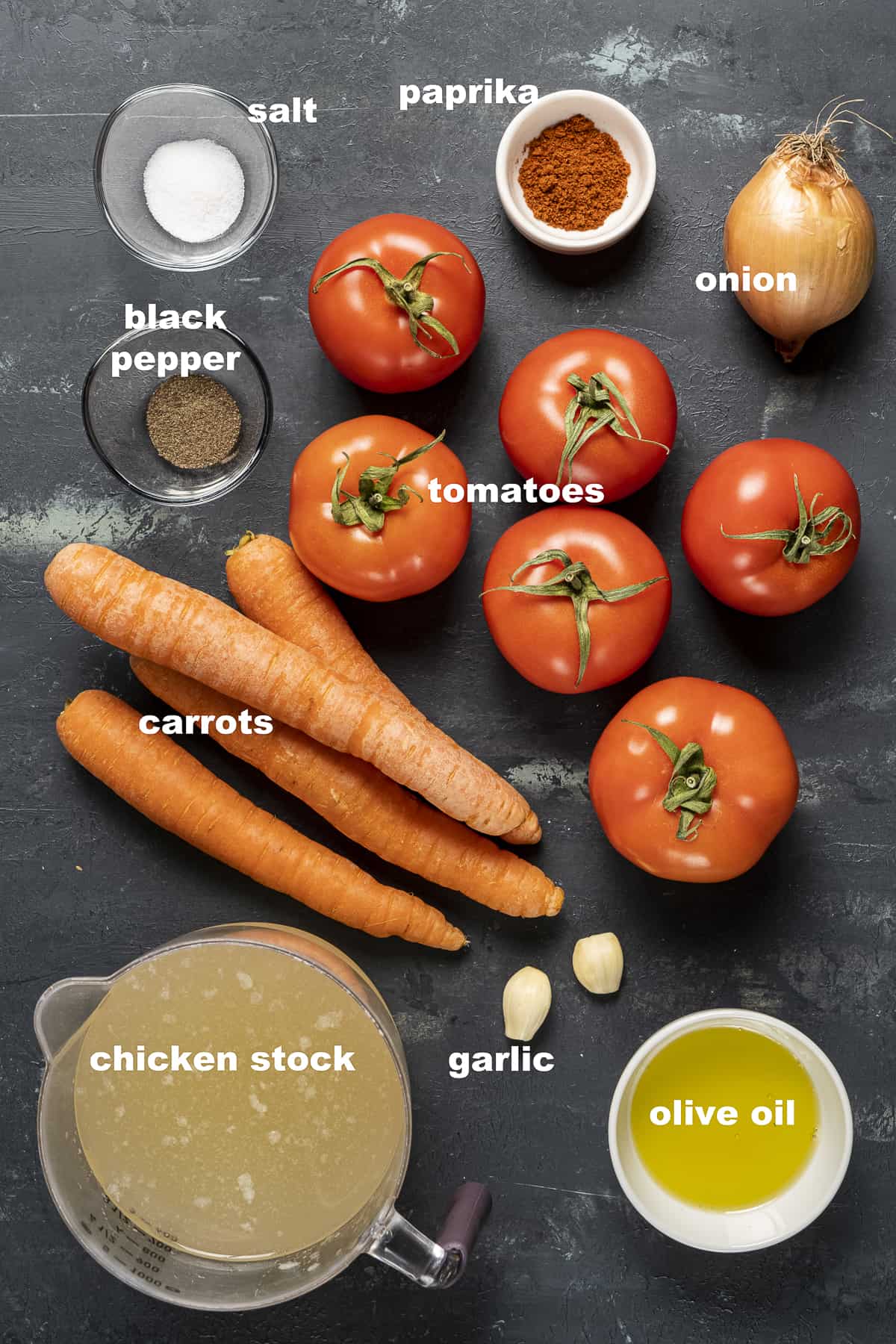 Tomatoes, carrots, chicken stock, olive oil, garlic, onion, salt, pepper and paprika on a dark background.