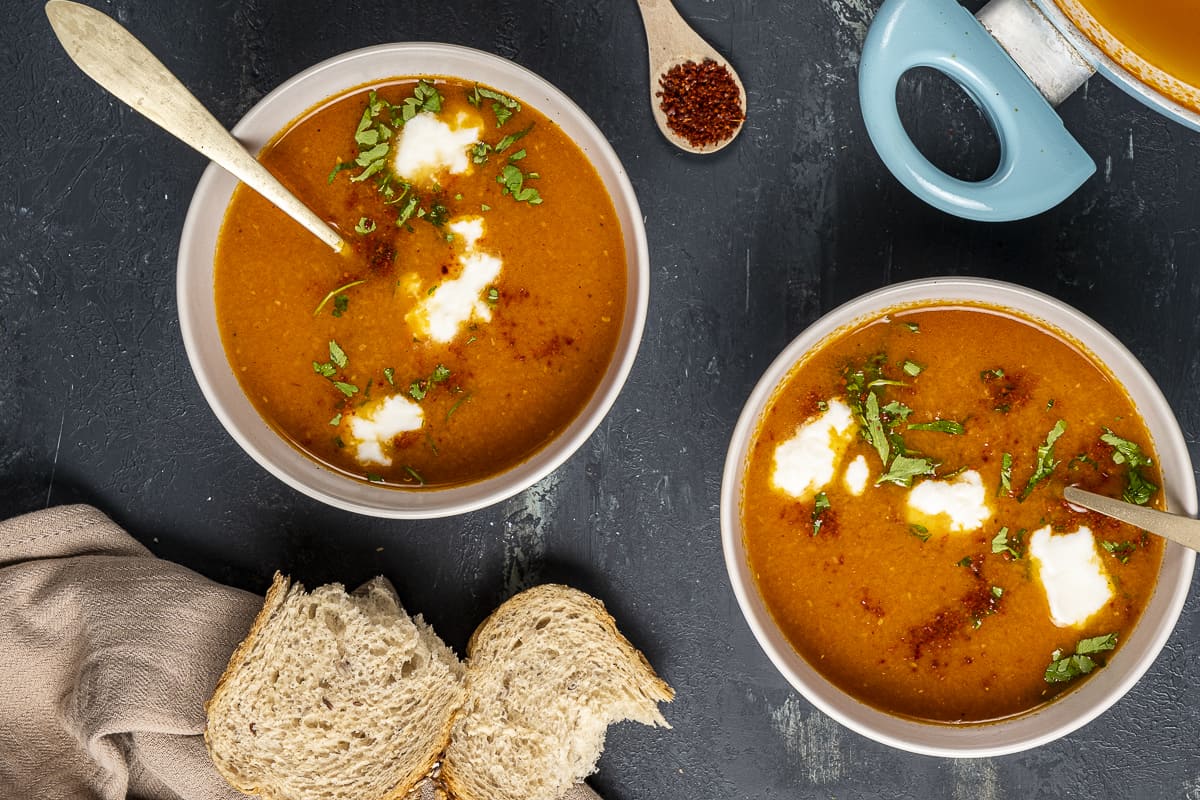 Tomato soup served in two bowls, garnished with yogurt and parsley and a spoon in each. Sliced bread and red pepper flakes on the side.
