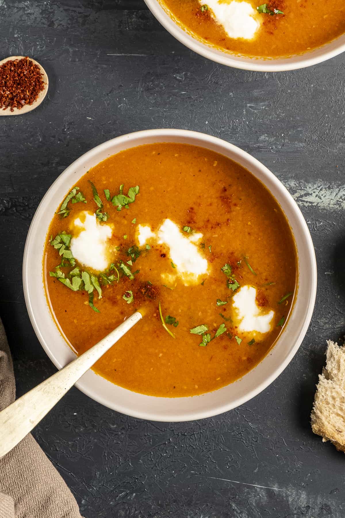 Tomato and carrot soup garnished with yogurt, parsley and red pepper flakes in a white bowl and a spoon inside it.