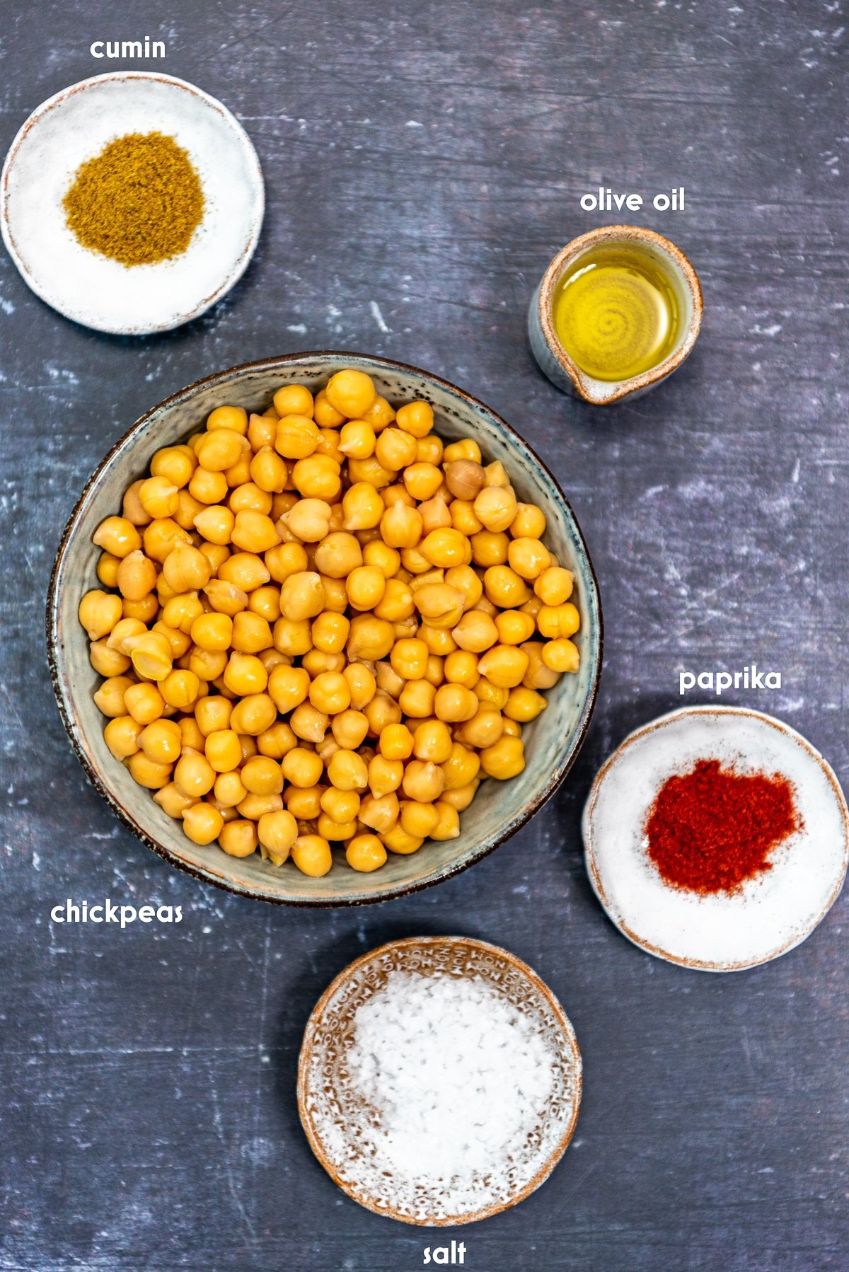 Cooked chickpeas, salt, paprika, cumin, salt and olive oil in separate bowls on a dark background.