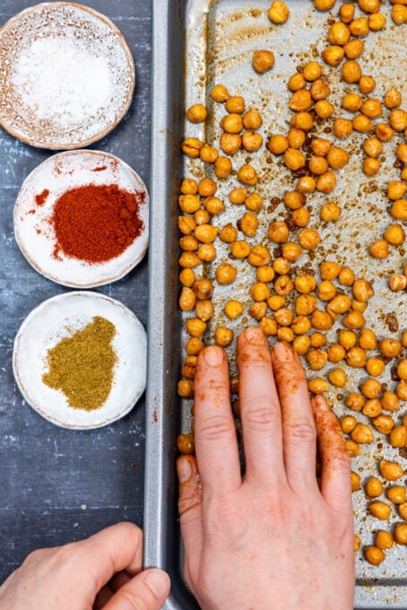 A hand coating the half roasted chickpeas with olive oil and spices on a baking sheet.