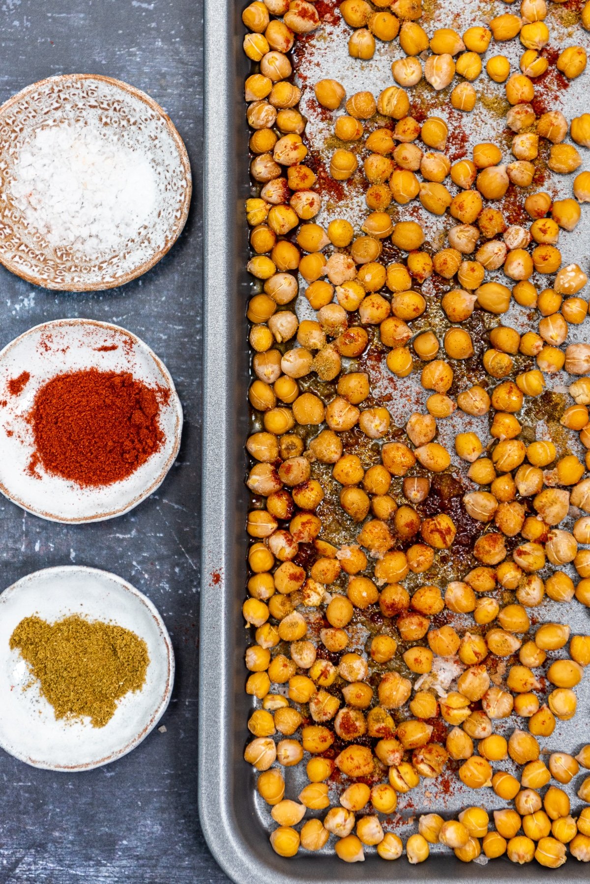 Roasted chickpeas on a baking sheet, a little spices on them. Paprika, cumin and salt in mini bowls on the side.