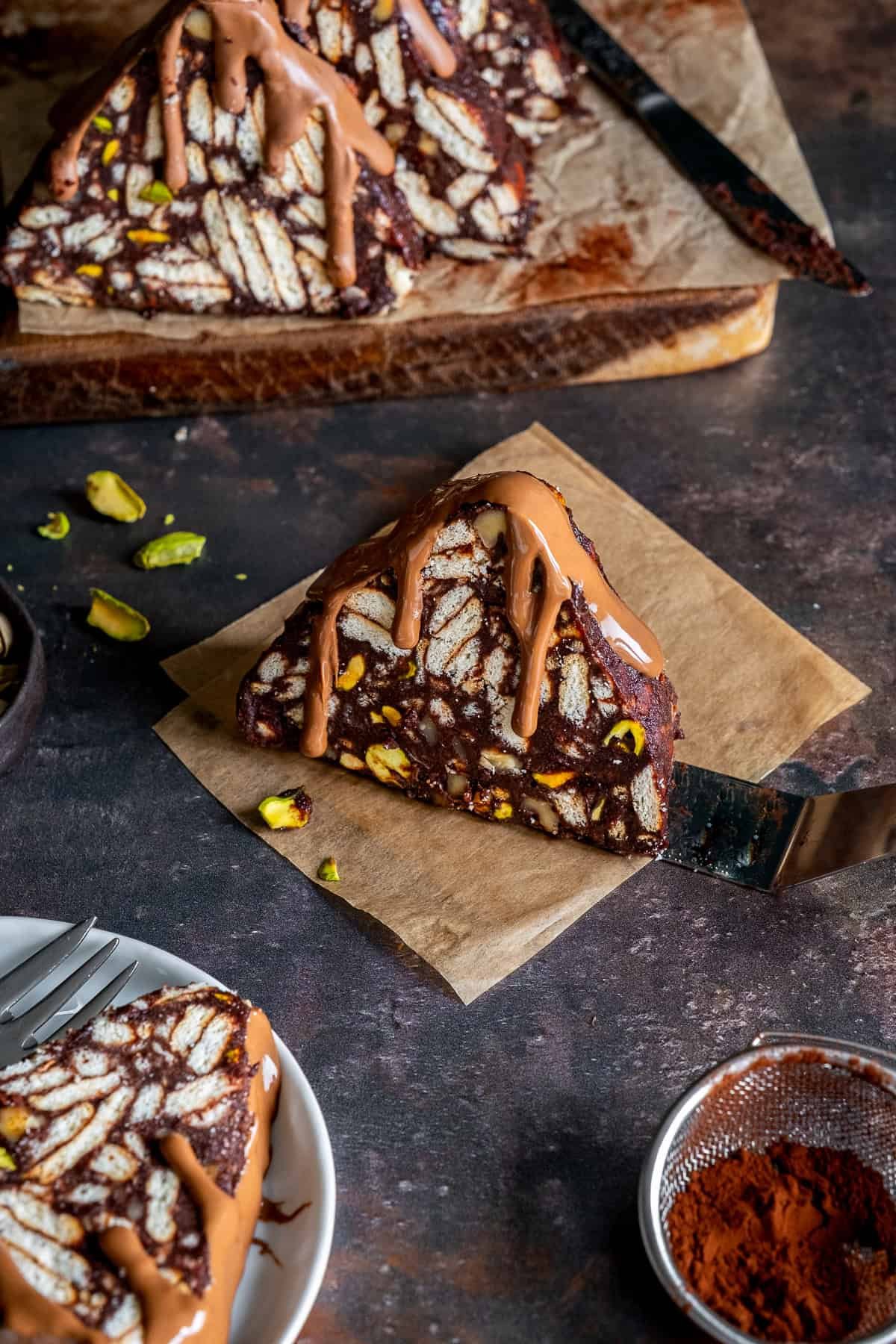 Slice of Turkish Mosaic Cake with melted chocolate on top, served on parchment paper with scattered pistachios and cocoa powder.