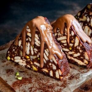 Triangular slices of Turkish Mosaic Cake topped with melted milk chocolate, on a wooden board with cocoa powder and pistachio pieces.