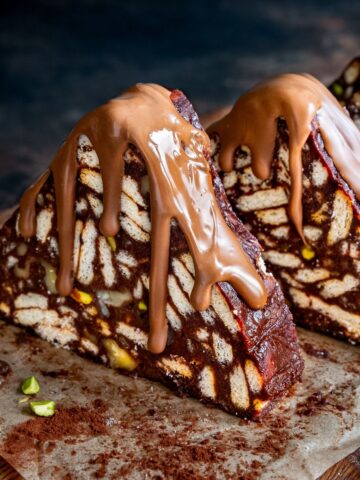 Triangular slices of Turkish Mosaic Cake topped with melted milk chocolate, on a wooden board with cocoa powder and pistachio pieces.