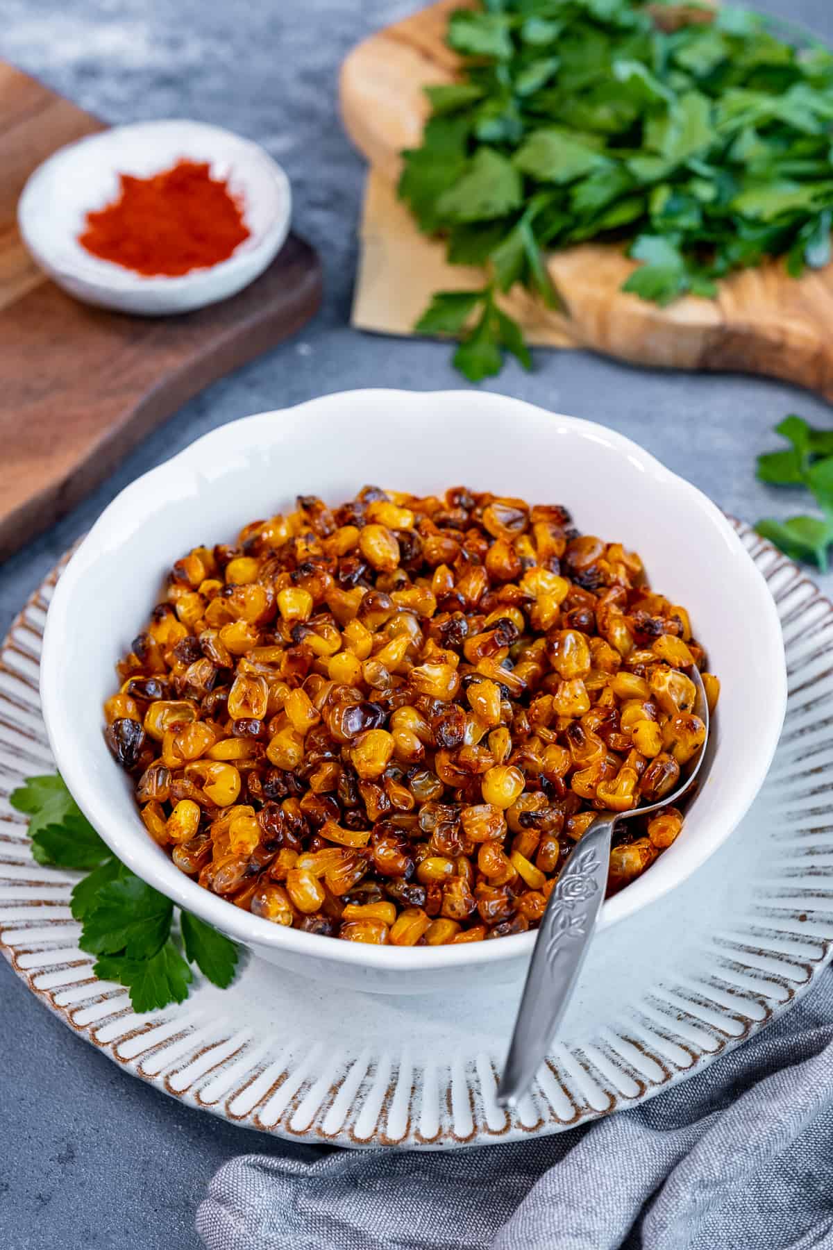 Roasted corn kernels in a white bowl. a bunch of parsley and paprika behind it.