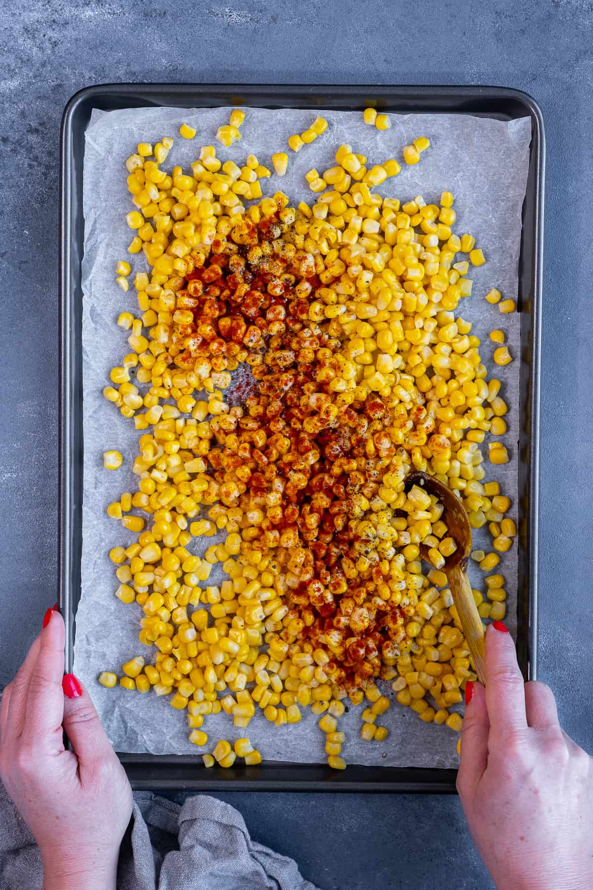 Hands mixing sweet corn kernels with spices on a baking sheet.