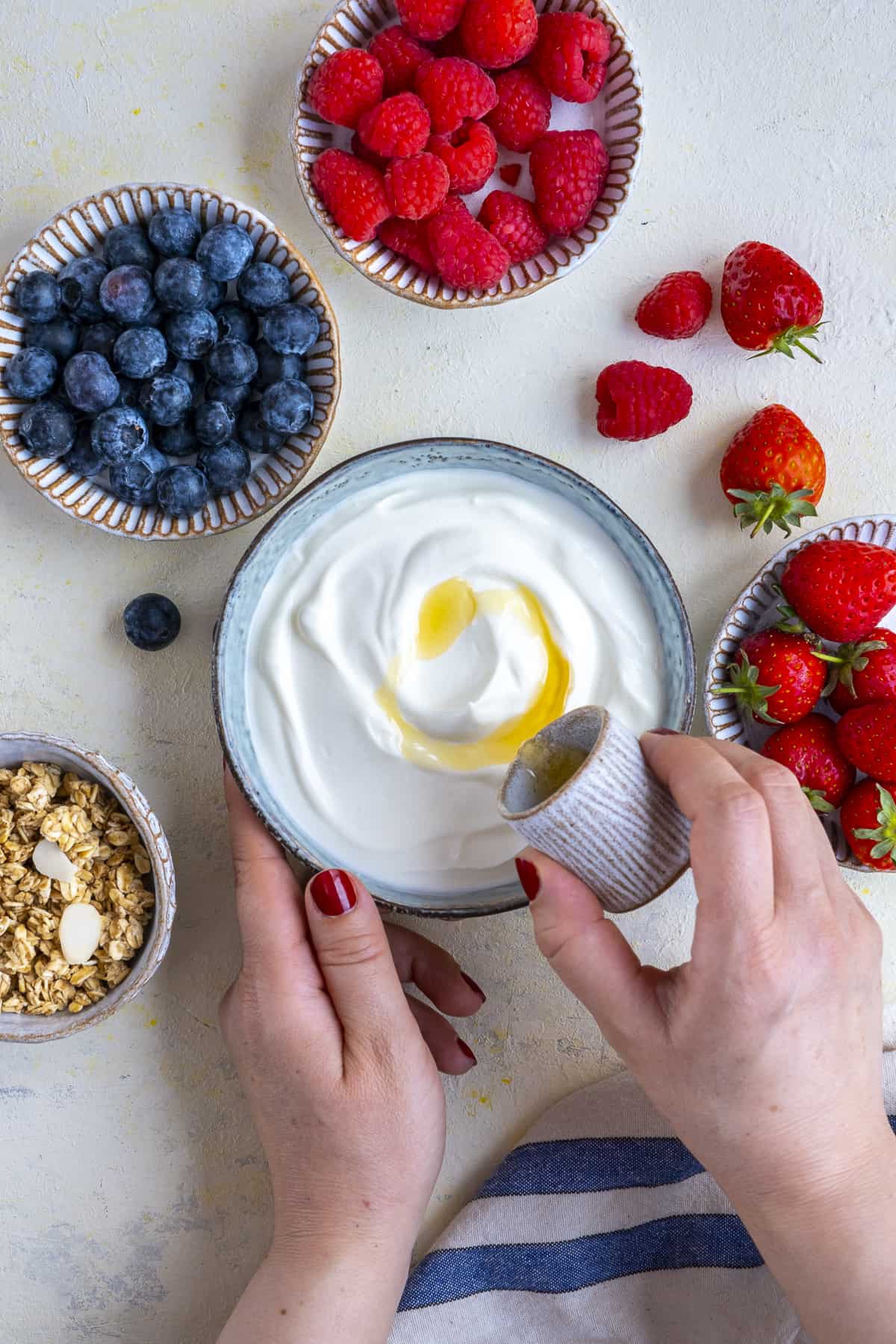 Hands pouring honey on yogurt in a bowl and berries on the side.