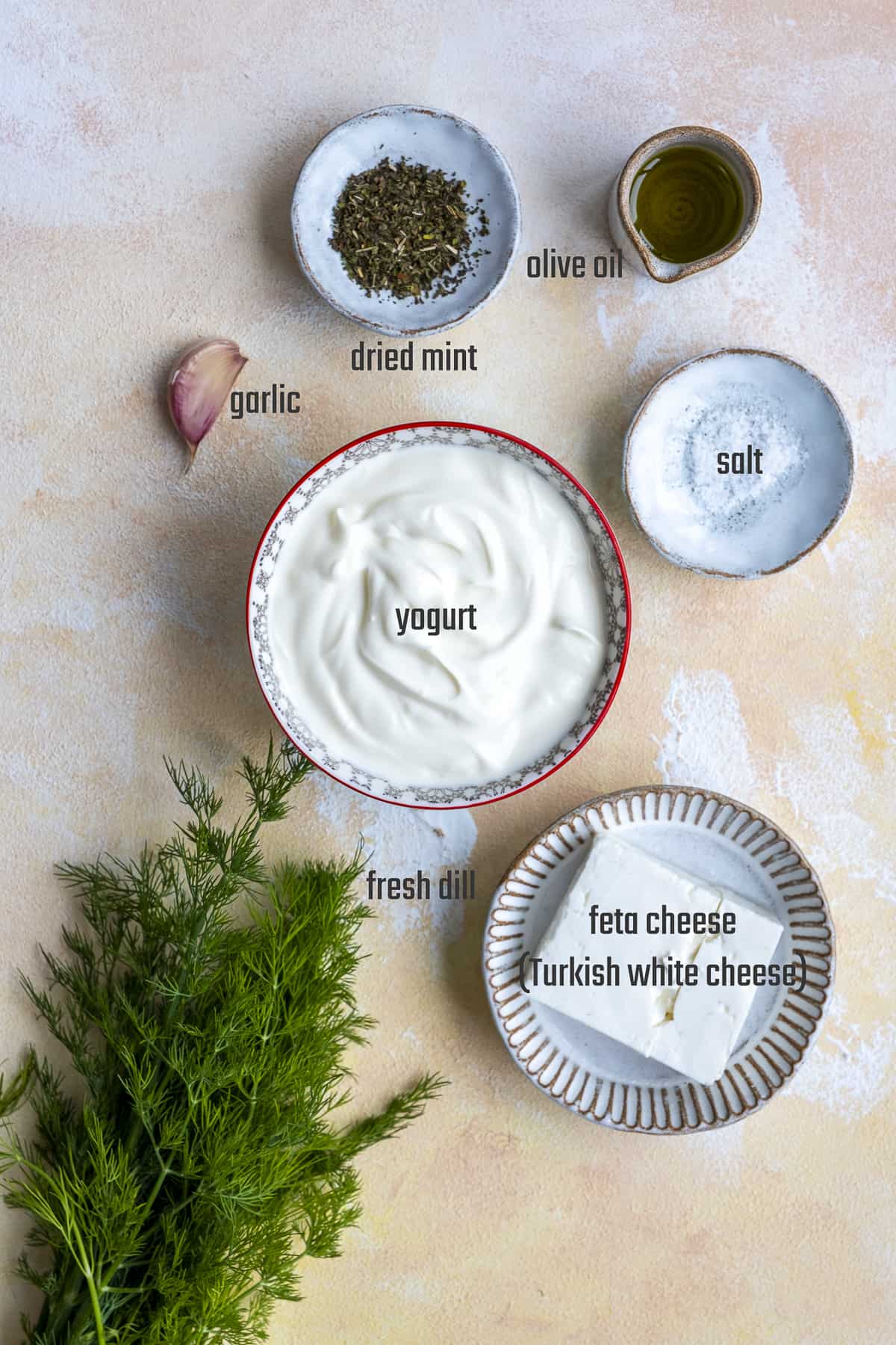 Greek yogurt, feta cheese, a clove of garlic, dried mint, olive oil, salt and fresh dill on a light background.