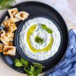 Haydari topped with olive oil, fresh dill and mint leaves served on a black plate and pita bread slices on the side.