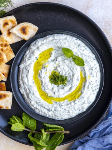 Haydari topped with olive oil, fresh dill and mint leaves served on a black plate and pita bread slices on the side.