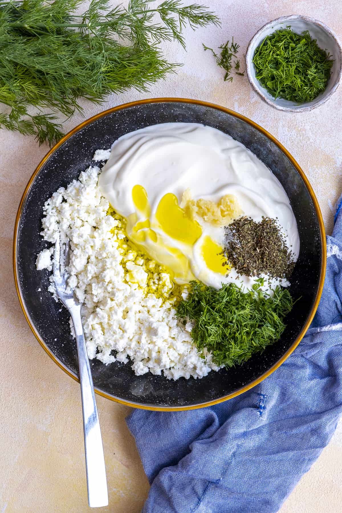 Mashed feta cheese, yogurt, olive oil, mashed garlic, chopped fresh dill and dried mint in a black bowl and a fork inside it.