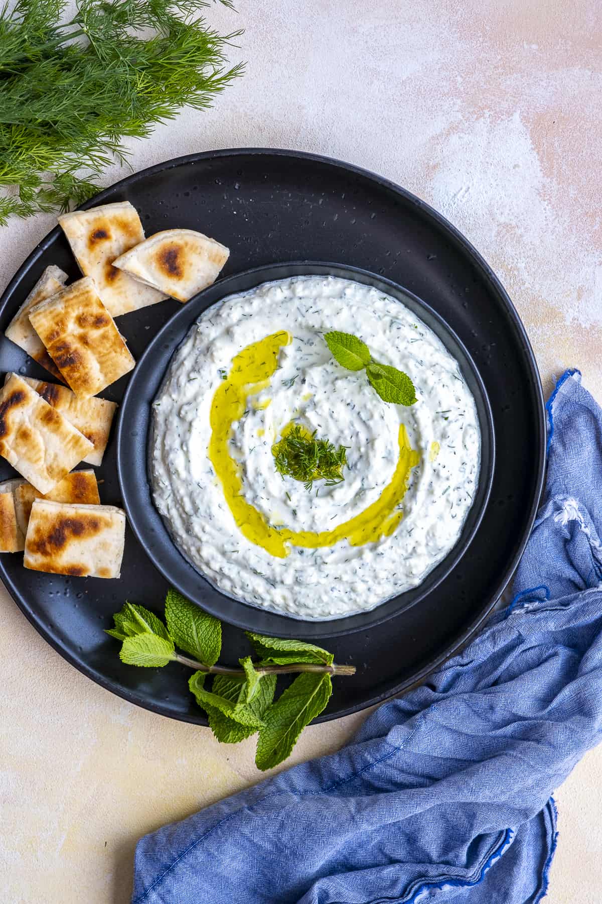 Haydari meze garnished with olive oil, fresh dill and mint leaves served on a black plate and pita bread slices on the side.