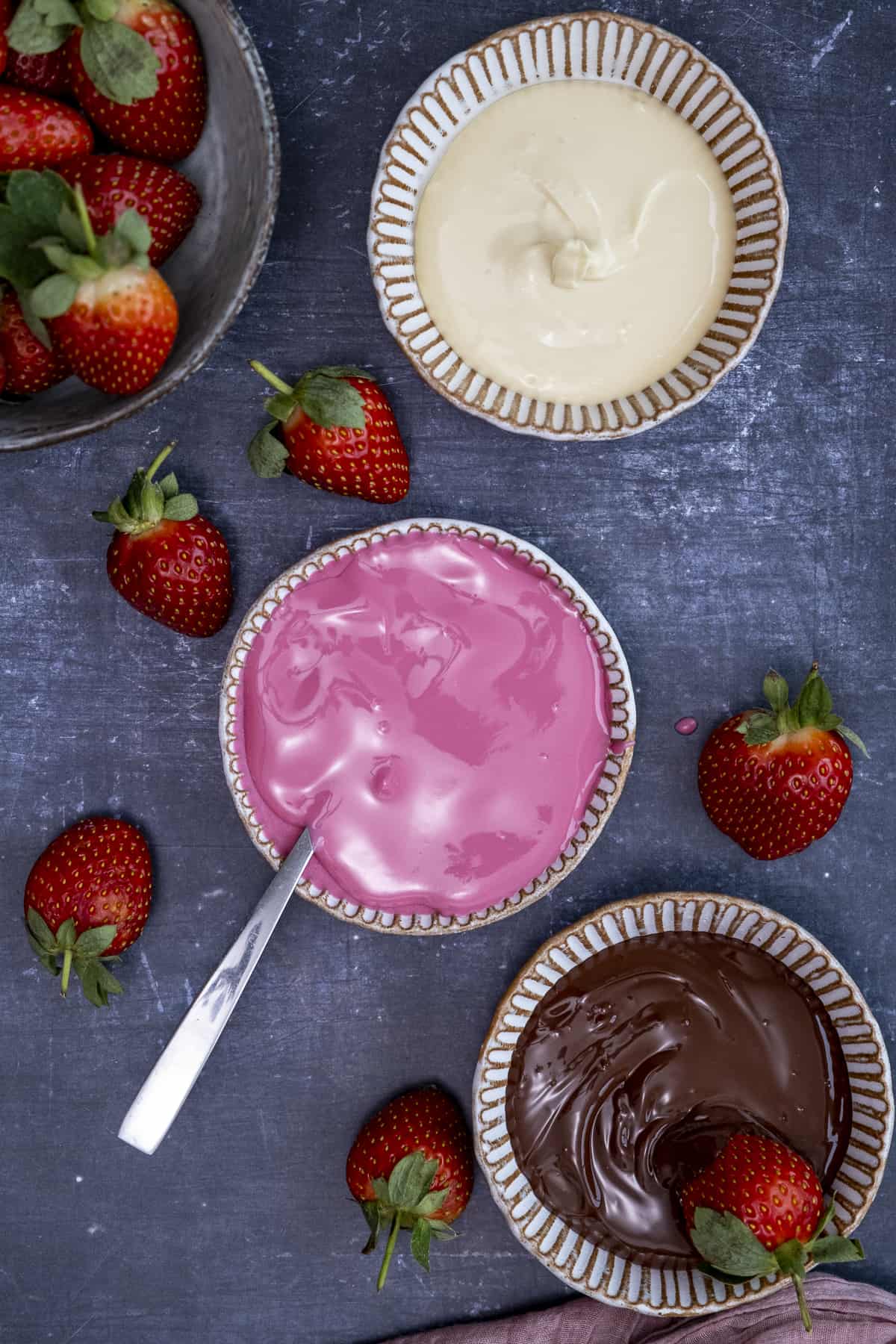 Melted white chocolate, pink candy melts and dark chocolate in separate bowls, strawberries around them on a dark background.