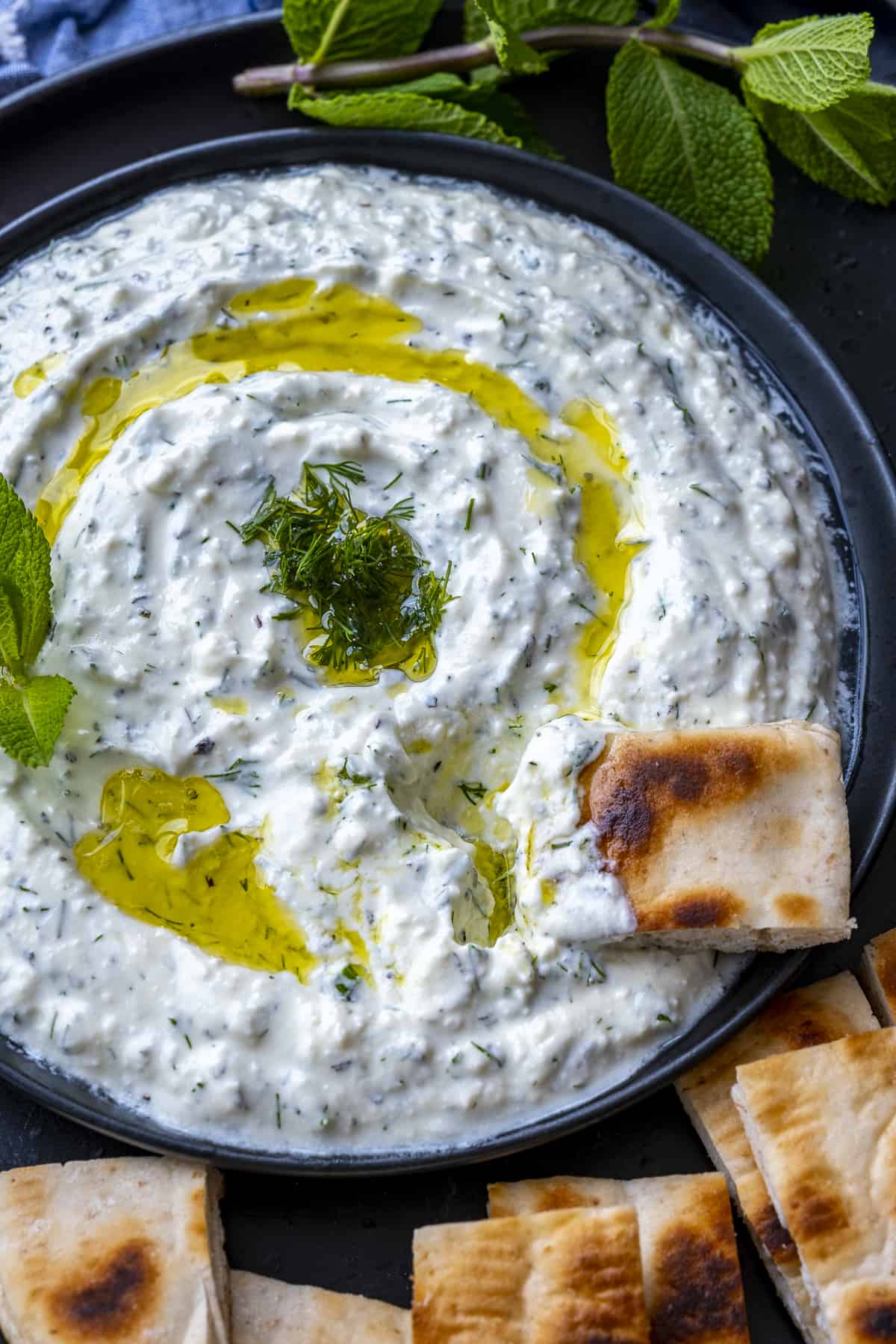 Haydari dip garnished with olive oil and fresh dill on a black plate. A slice of pita bread dipped into it.