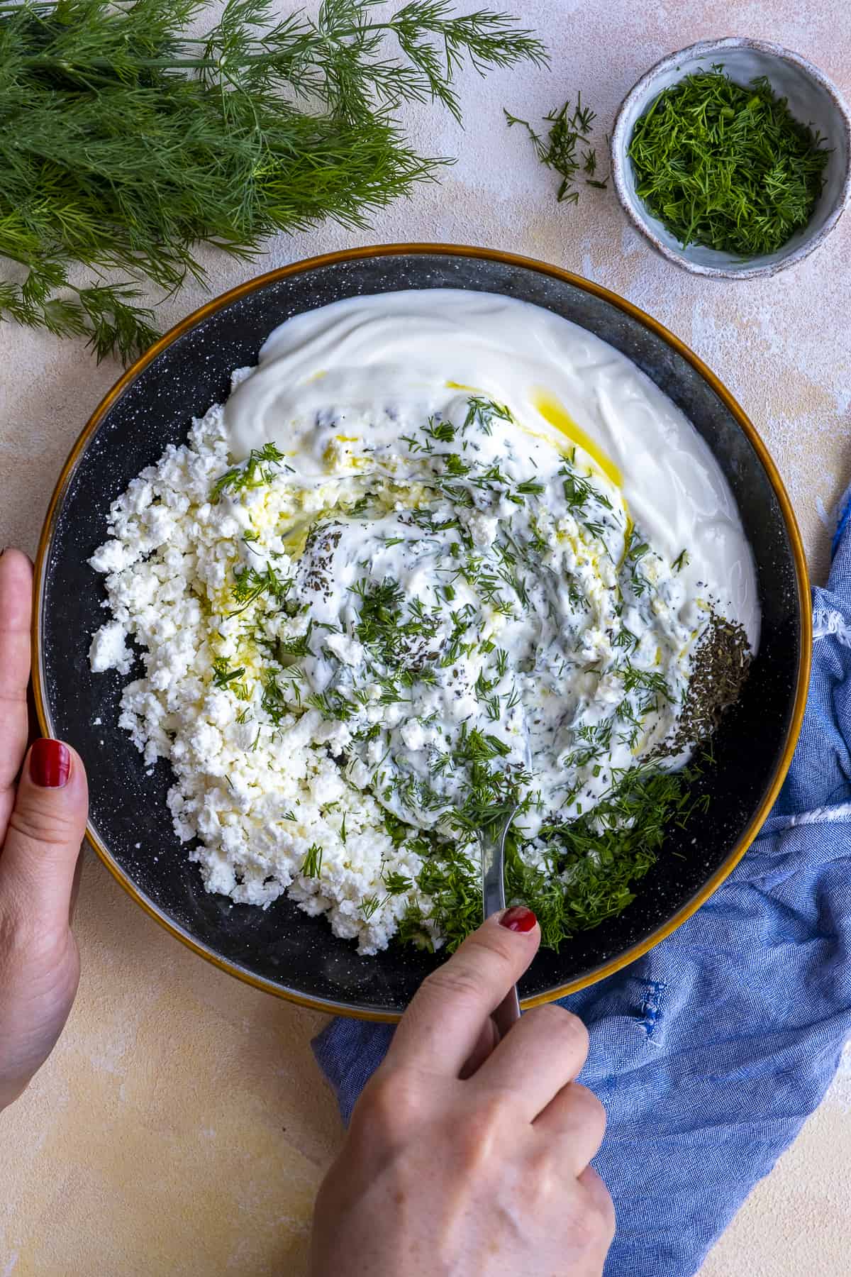 Hands stirring yogurt, feta cheese, fresh dill, dried mint and olive oil in a black bowl.