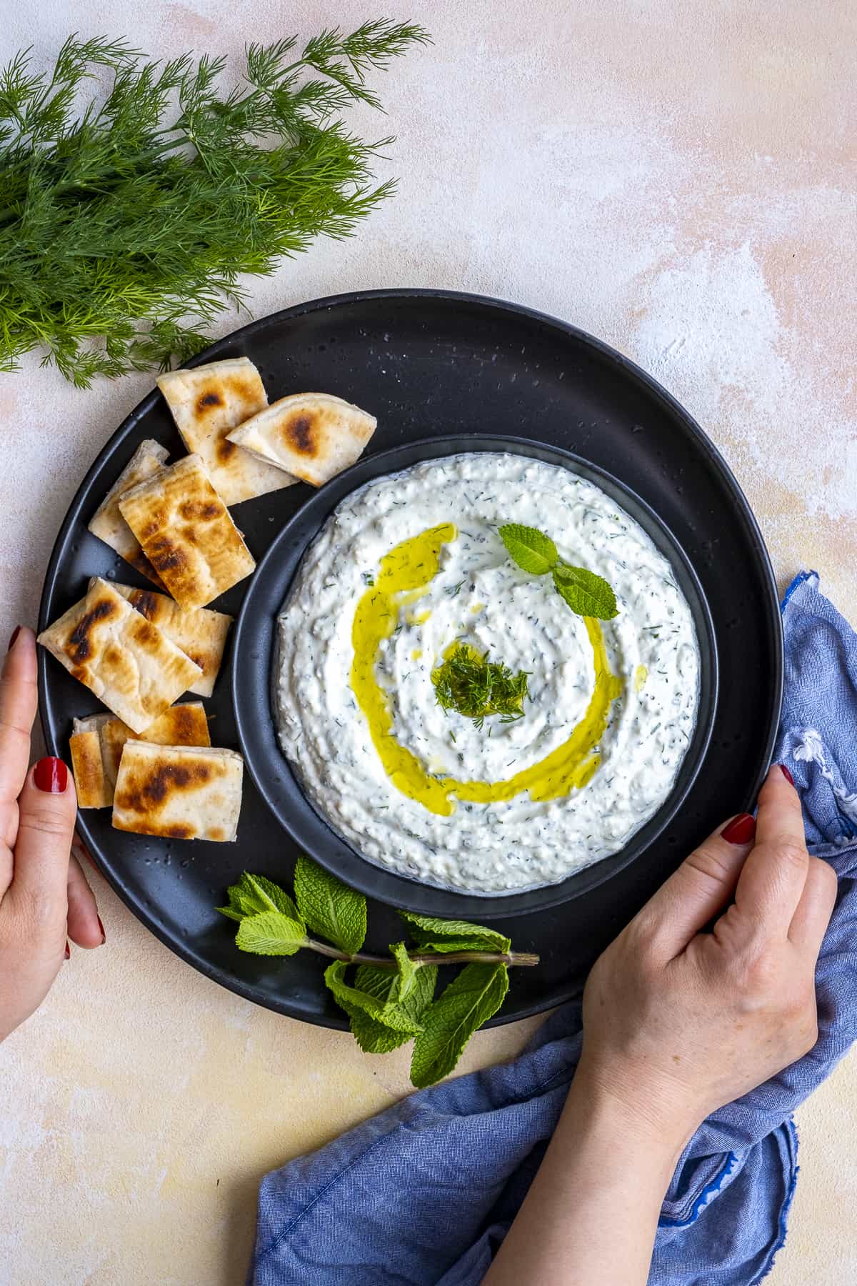 Woman hands holding haydari meze and pita bread slices on a black plate. Fresh dill and mint leaves on the side.