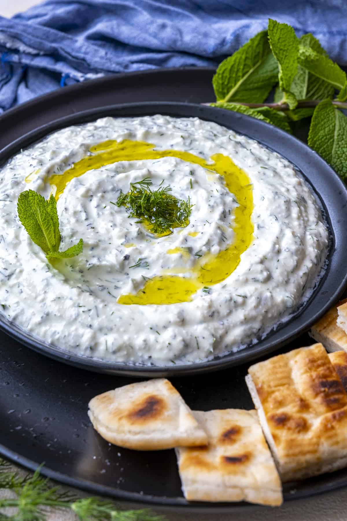 Haydari garnished with olive oil and fresh herbs accompanied by pita bread slices on a black plate.