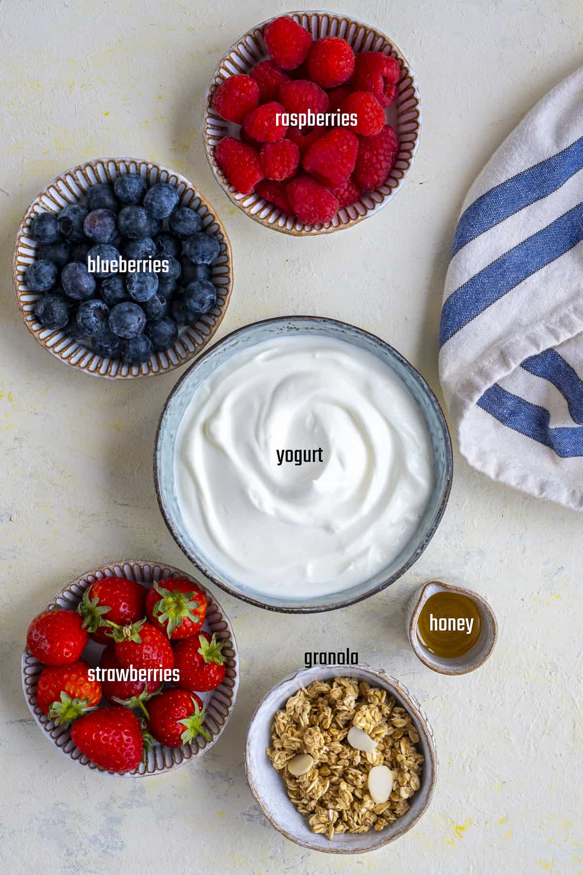 Yogurt, raspberries, blueberries, strawberries, granola and honey in separate bowls on a light background.