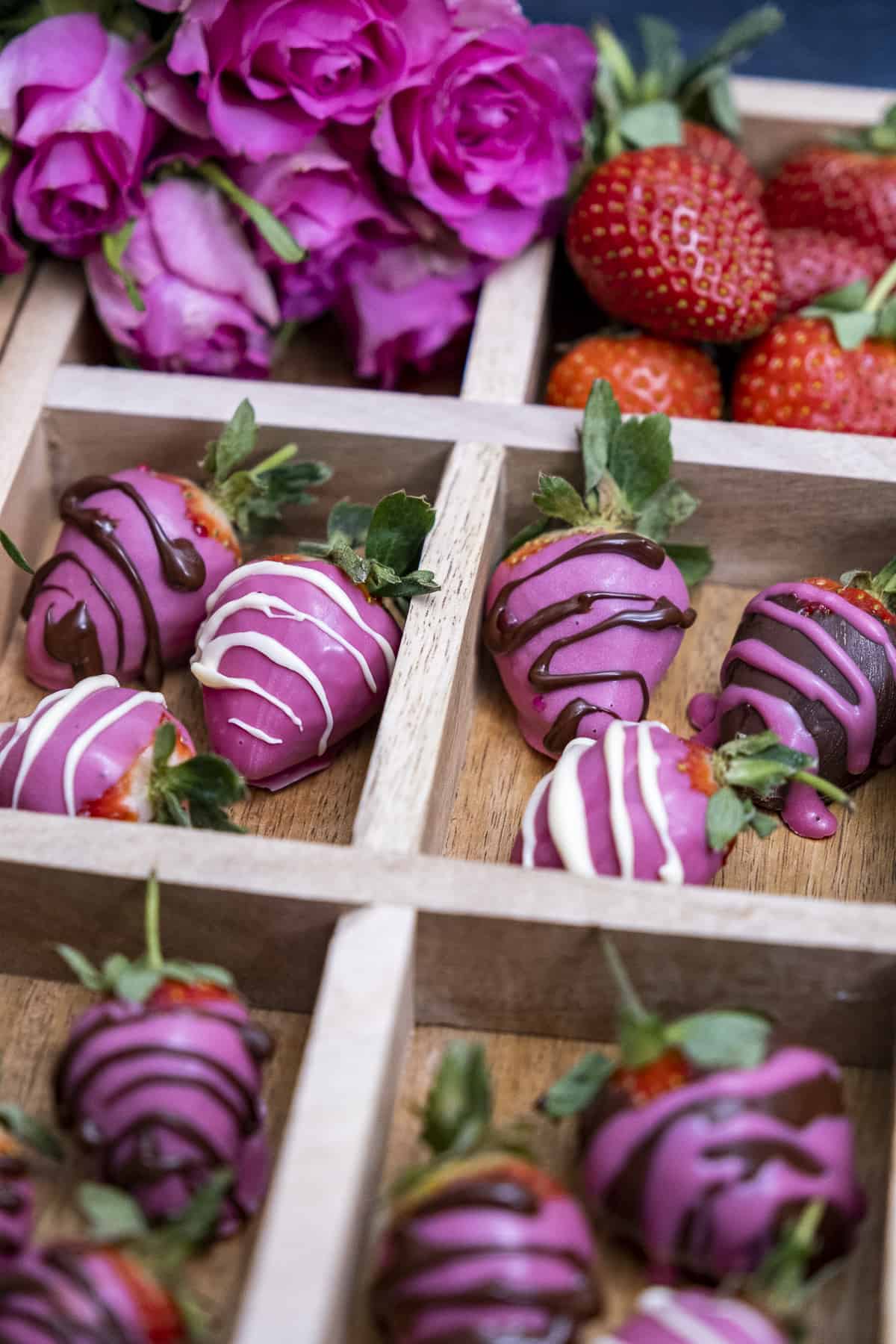 Pink chocolate strawberries in a wooden box with sections, pink roses and strawberries behind these.