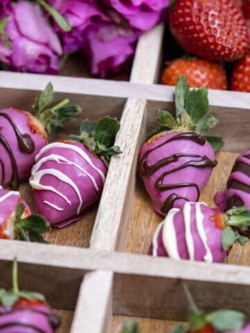 Pink chocolate strawberries in a wooden box with sections, pink roses and fresh strawberries behind them.