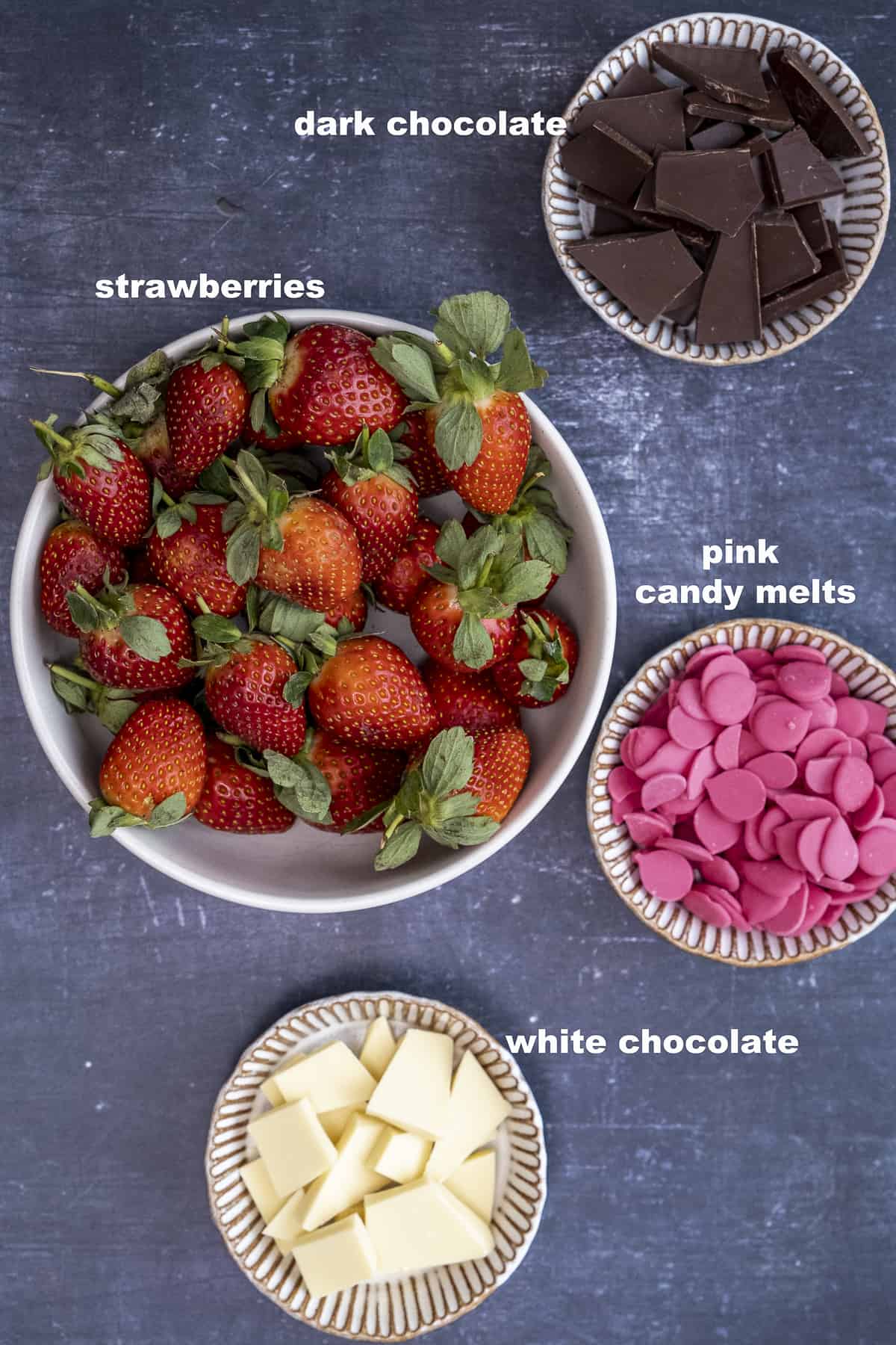 Strawberries, chopped dark chocolate, pink candy melts and chopped white chocolate in separate bowls on a dark background.