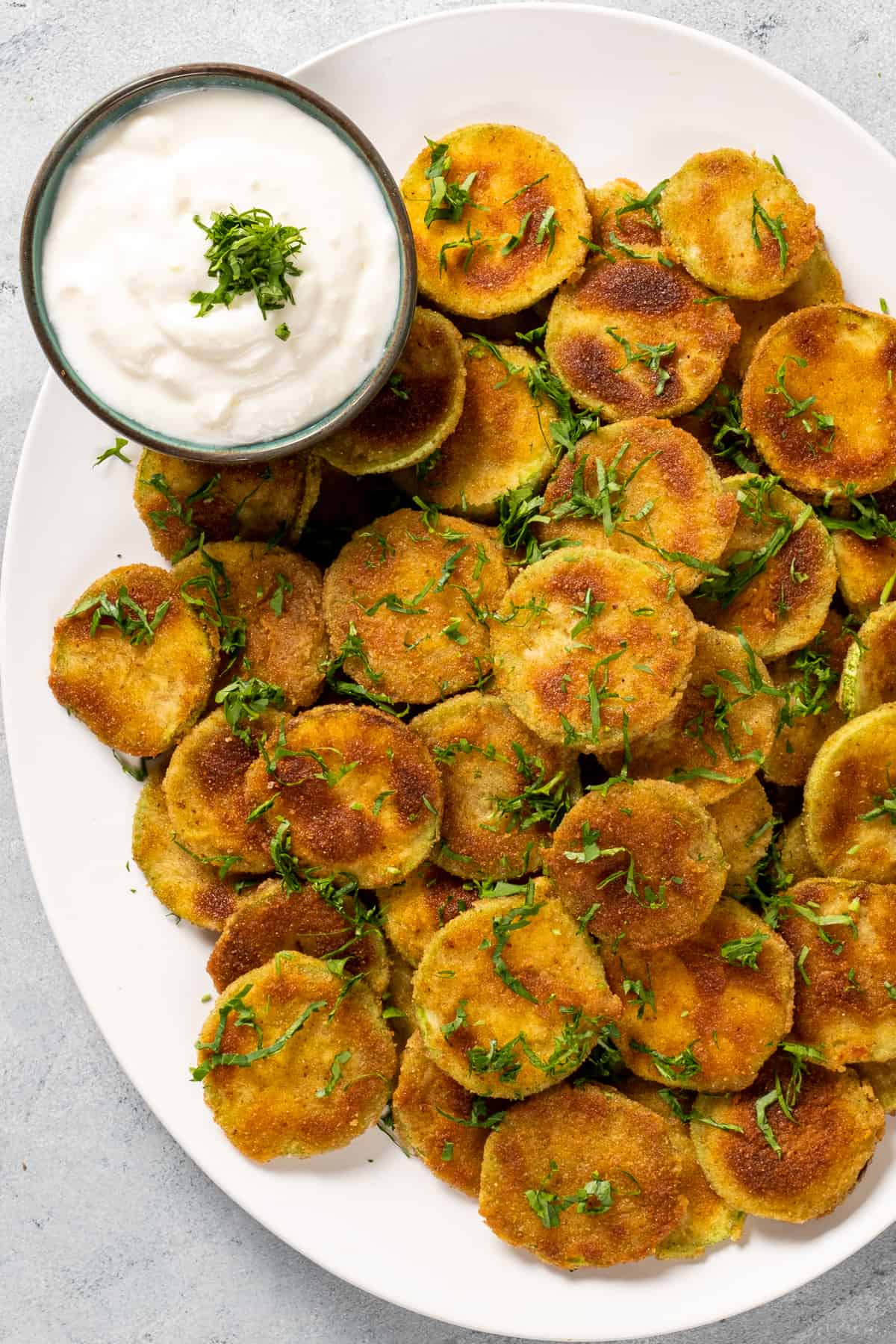 Fried zucchini chips served on a white oval dish and a small bowl of yogurt sauce on the side.