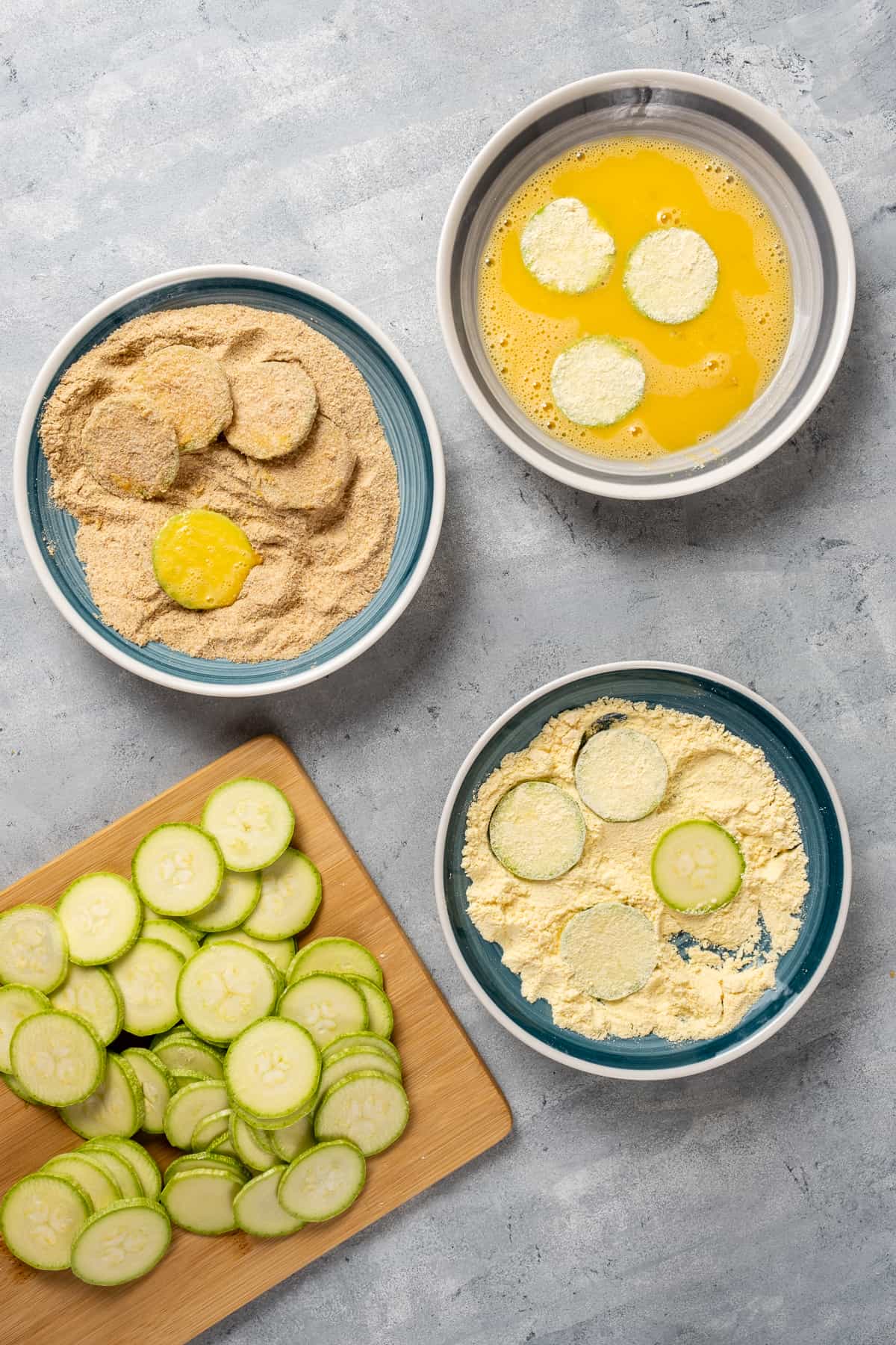 Cornmeal, breadcrumbs, beaten eggs in separate bowls and zucchini slices on a wooden board on the side.