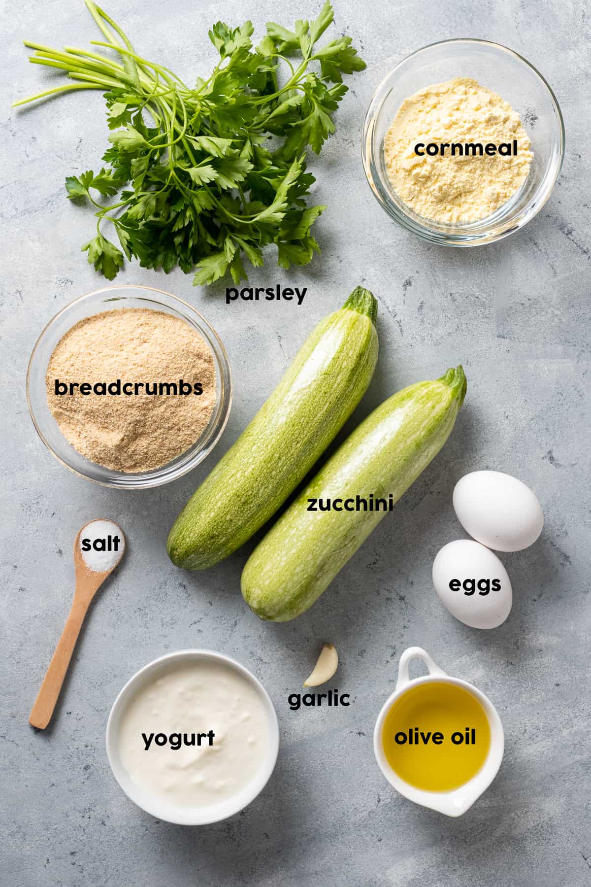 Zucchini, eggs, cornmeal, parsley, olive oil, garlic, salt and pepper on a grey background.