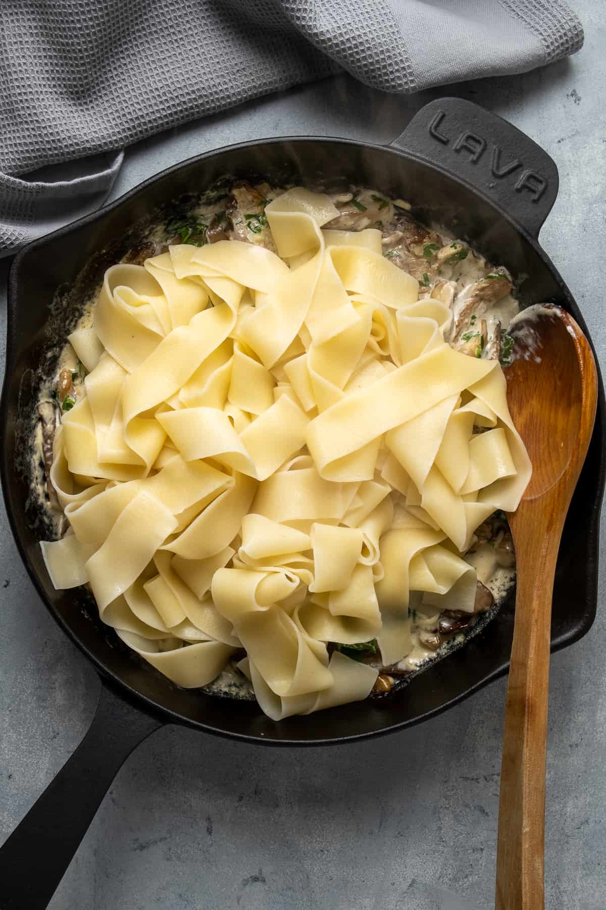 Cooked pasta combining with creamy mushroom sauce in a cast iron skillet and a wooden spoon inside it.