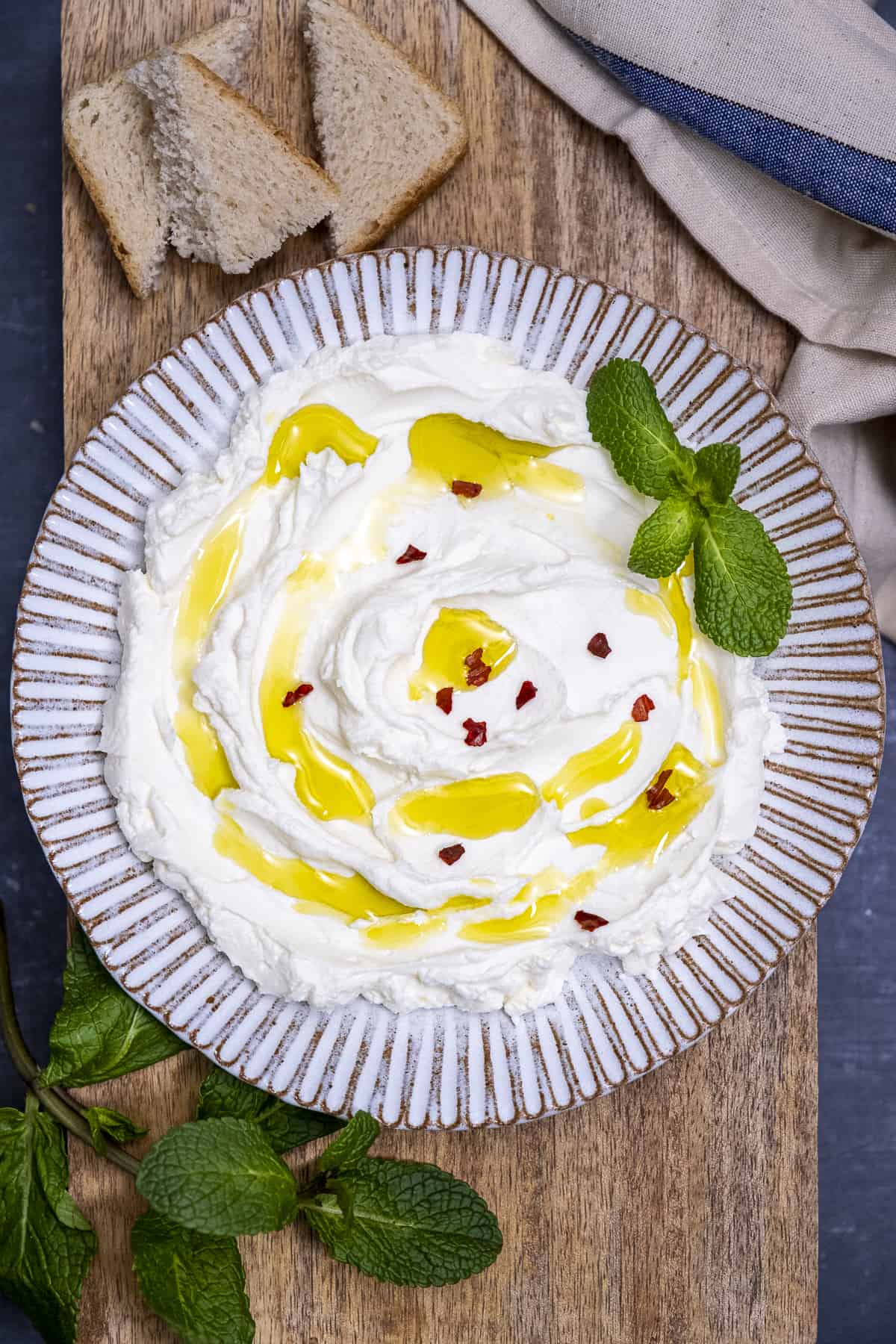 Strained thick yogurt drizzled with olive oil and garnished with fresh mint leaves and red pepper flakes on a white ceramic plate, small slices of bread on the side.