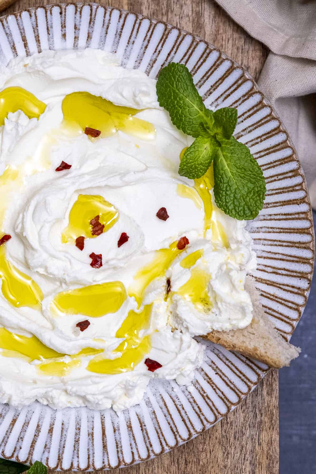 Greek yogurt topped with red pepper flakes and drizzled with olive oil, a sprig of fresh mint on one side and a little bread is dipped into it.
