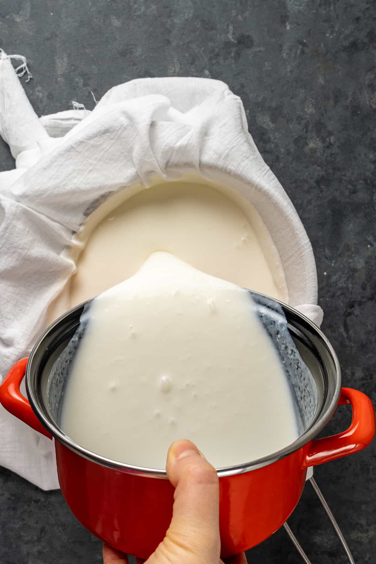 A hand pouring regular yogurt from a red pot onto a white muslin.