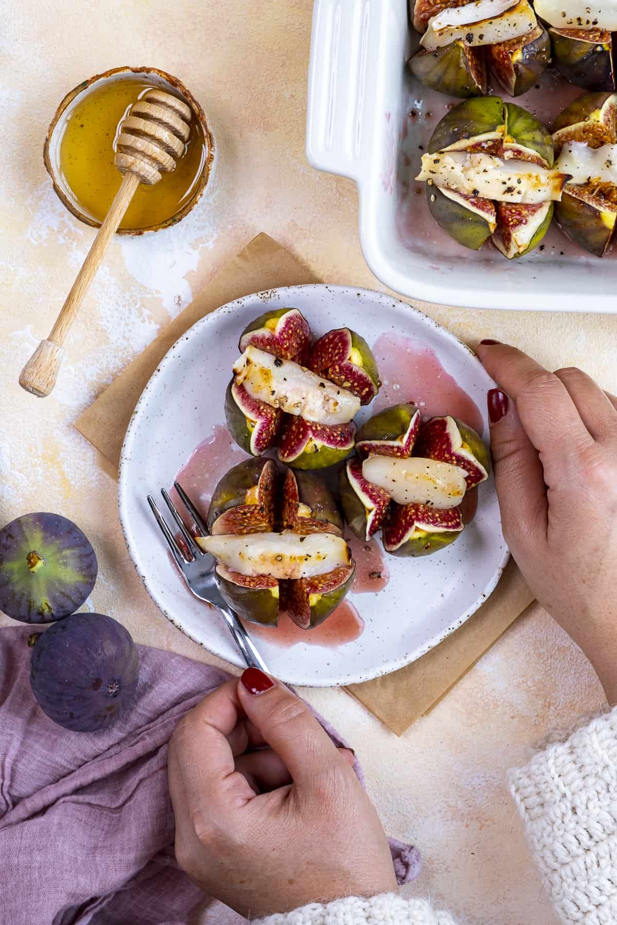 Woman hands holding a plate of baked figs with cheese, fresh figs and honey on the side.