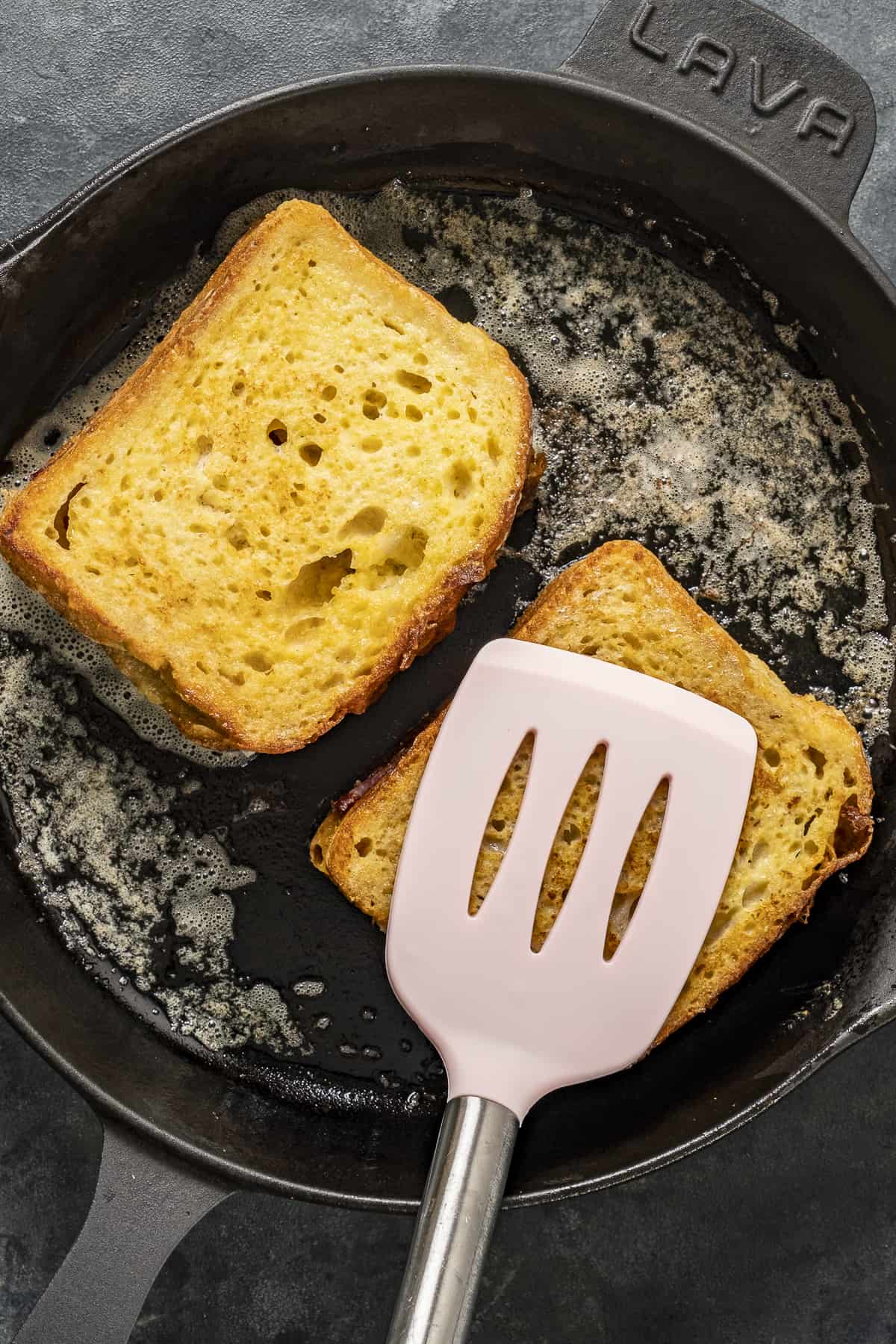 A spatula pressing on a slice of bread cooking in a pan.