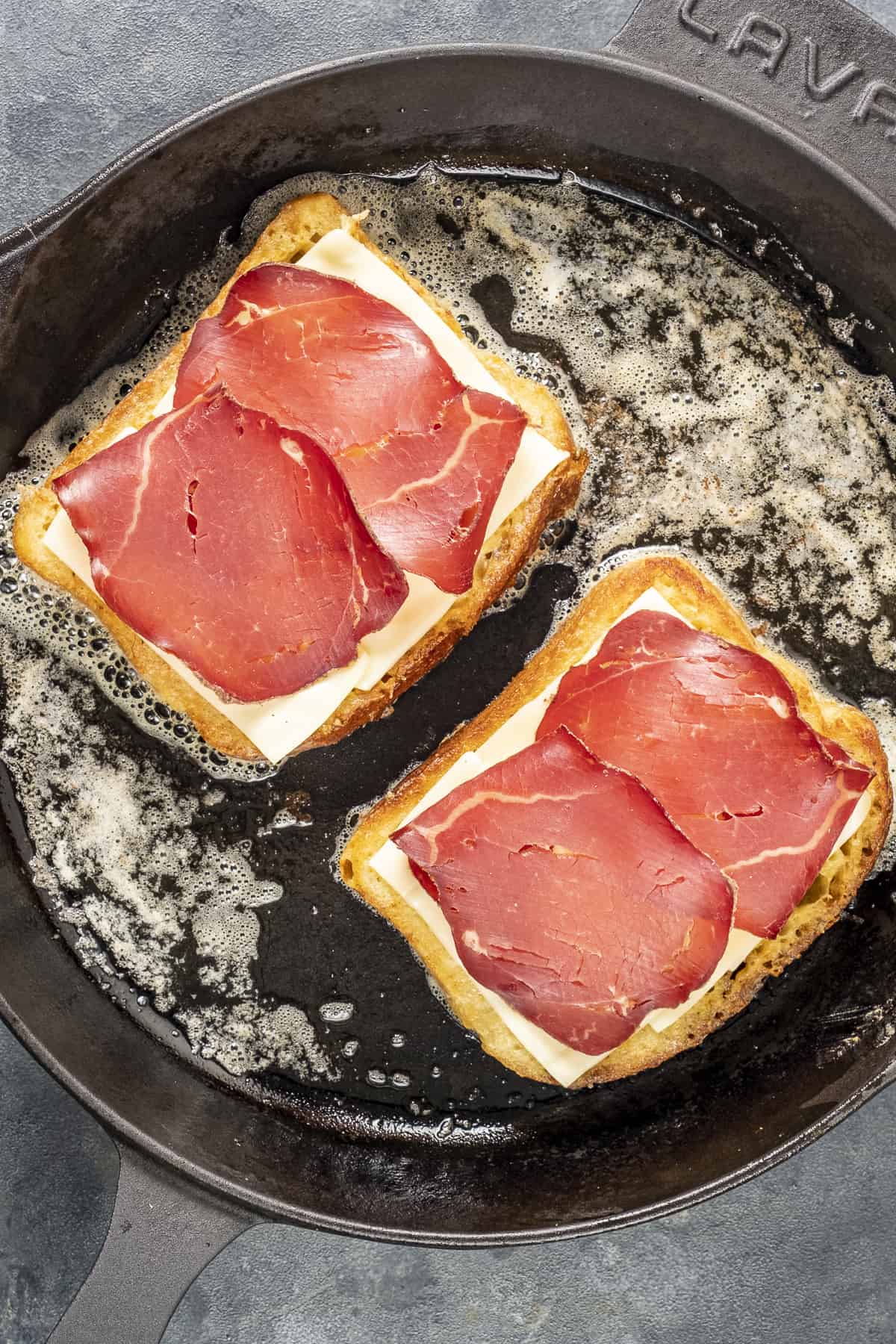 Bread slices topped with cheese and cold meat slices frying in a pan.