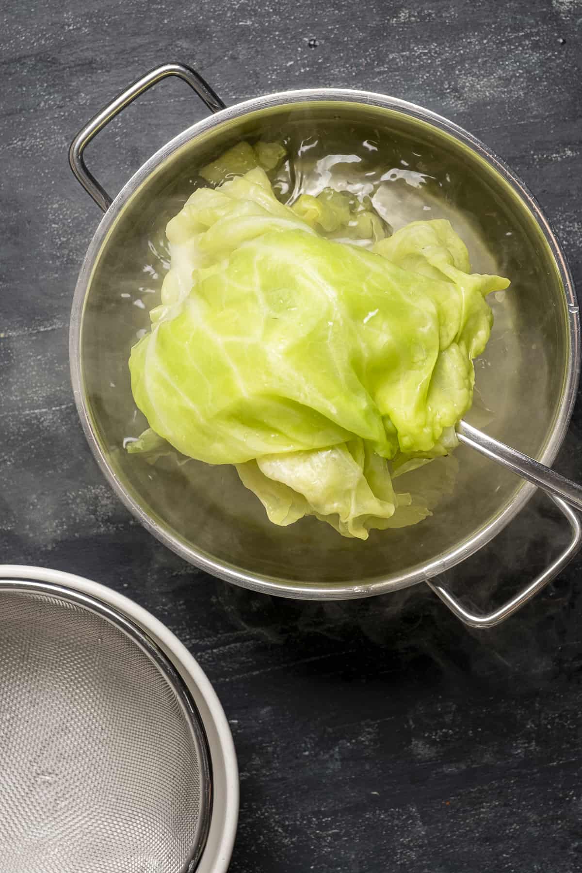 Cabbage leaves blanching in boiling water in a pot and a strainer on the side.