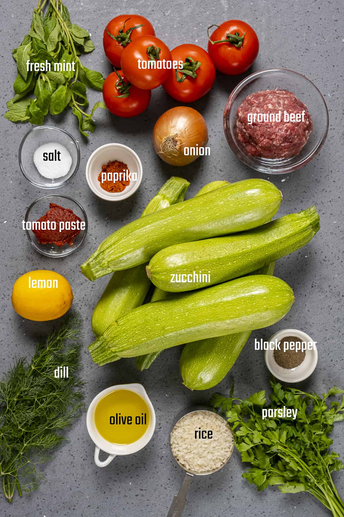 Zucchini, tomatoes, parsley, dill, mint, rice, ground beef, onion, tomato paste, olive oil and spices on a grey background.